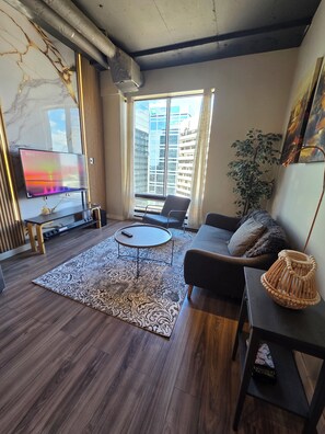 A modern living room with a marble wall, a flat-screen TV, a sofa, a coffee table, and a large window with a city view. - Cozy 16th-Floor Downtown Loft | City Views | Steps to ICE District (Edmonton)