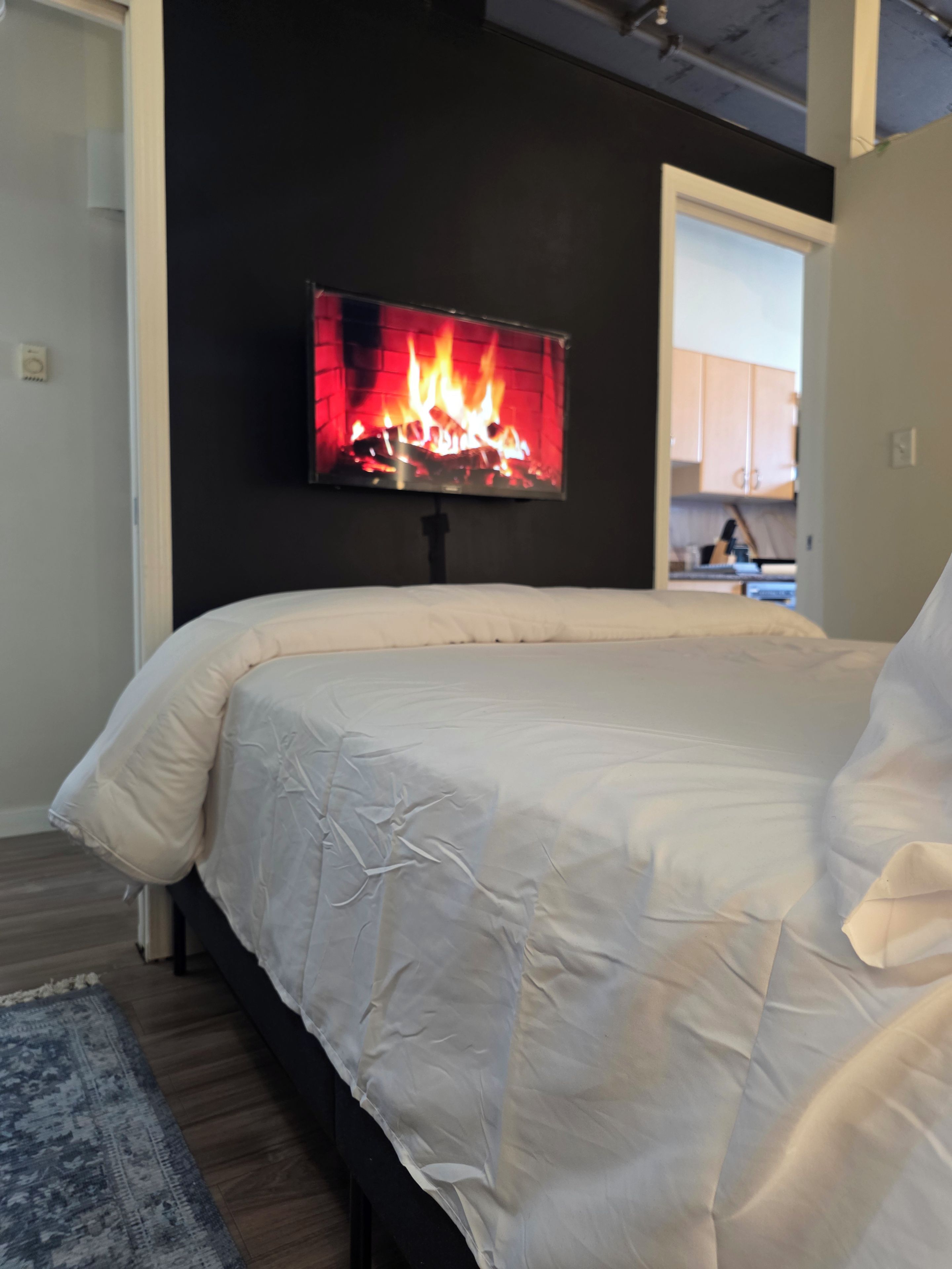 A bedroom with a bed, a flat-screen TV mounted on the wall, and a visible kitchen area in the background.