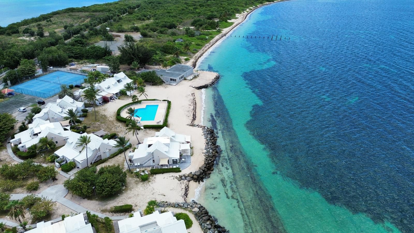 An aerial view of a coastal resort with multiple white buildings, a swimming pool, and a rocky shoreline.