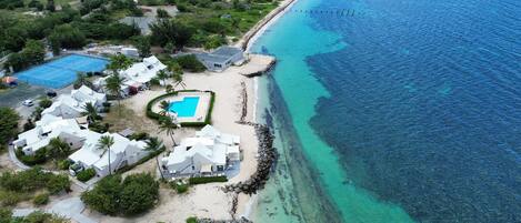 An aerial view of a coastal resort with multiple white buildings, a swimming pool, and a rocky shoreline.