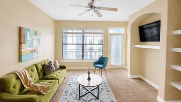 Un salon avec un canapé vert, un fauteuil bleu, une table basse ronde, un ventilateur de plafond et un téléviseur fixé au mur.