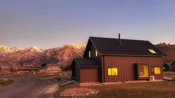 Une cabane en bois à deux étages, dotée d’une grande fenêtre et d’une cheminée, se dressant sur un fond de montagnes.