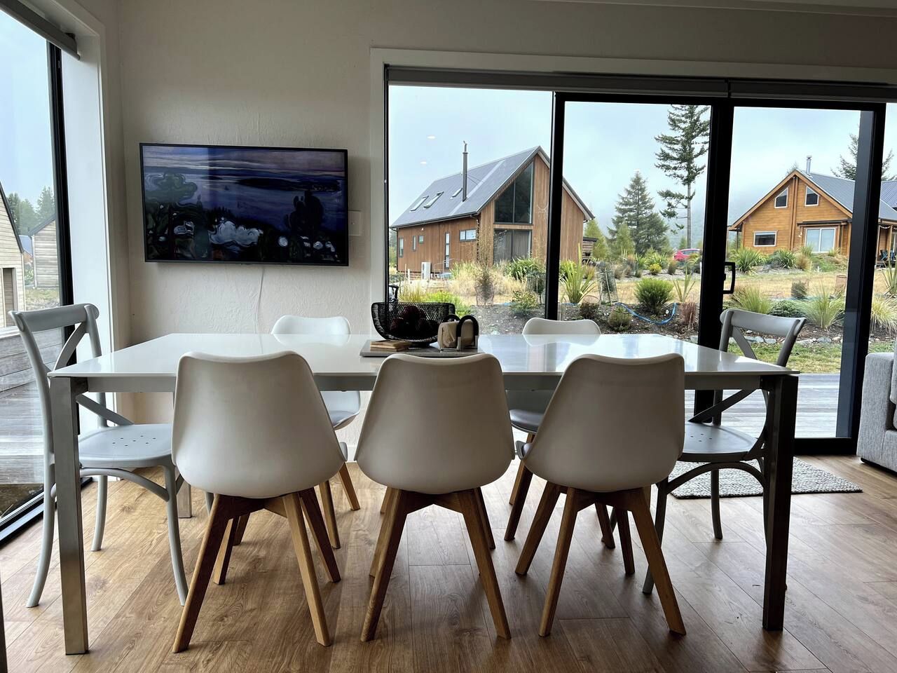 Un coin repas avec une table et des chaises, un téléviseur à écran plat fixé au mur et une vue sur les maisons à travers de grandes fenêtres.
