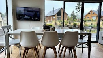 Un coin repas avec une table et des chaises, un téléviseur à écran plat fixé au mur et une vue sur les maisons à travers de grandes fenêtres.