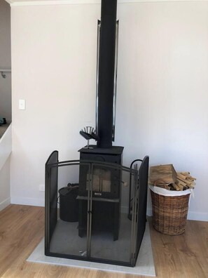 A wood-burning stove with a glass enclosure, a basket of firewood, and a staircase in the background.