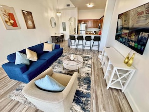 A modern living room with a blue sofa, a beige armchair, and a coffee table. There is a kitchen area with a bar and stools in the background. - Balcony Lake Views | Pool, Gym, WiFi & Parking (Orlando)