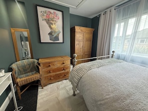 A bedroom with a bed, a wooden dresser, a wicker chair, a mirror, and a painting on the wall. - Beautiful House 5 Mins Walk Into Ulverston (Ulverston)