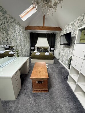 Ein gemütliches Schlafzimmer mit Dachfenster, einem Bett mit grüner Bettdecke, einer Holzkiste, einem Schreibtisch und einem Fernseher an der Wand.