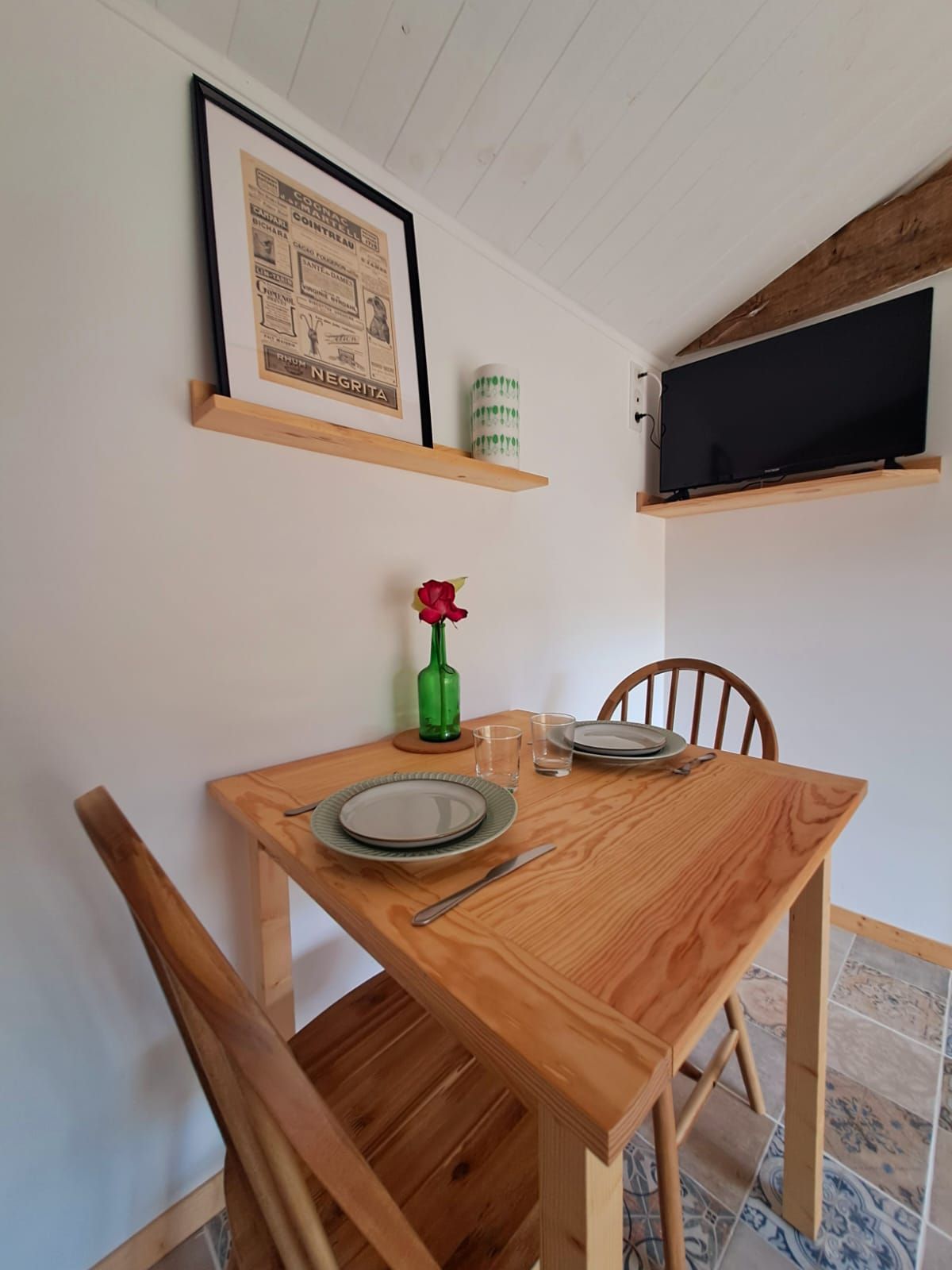 Una mesa de madera con platos, un cuchillo y una botella verde. Encima de la mesa, hay un cuadro enmarcado y un televisor montado en la pared.