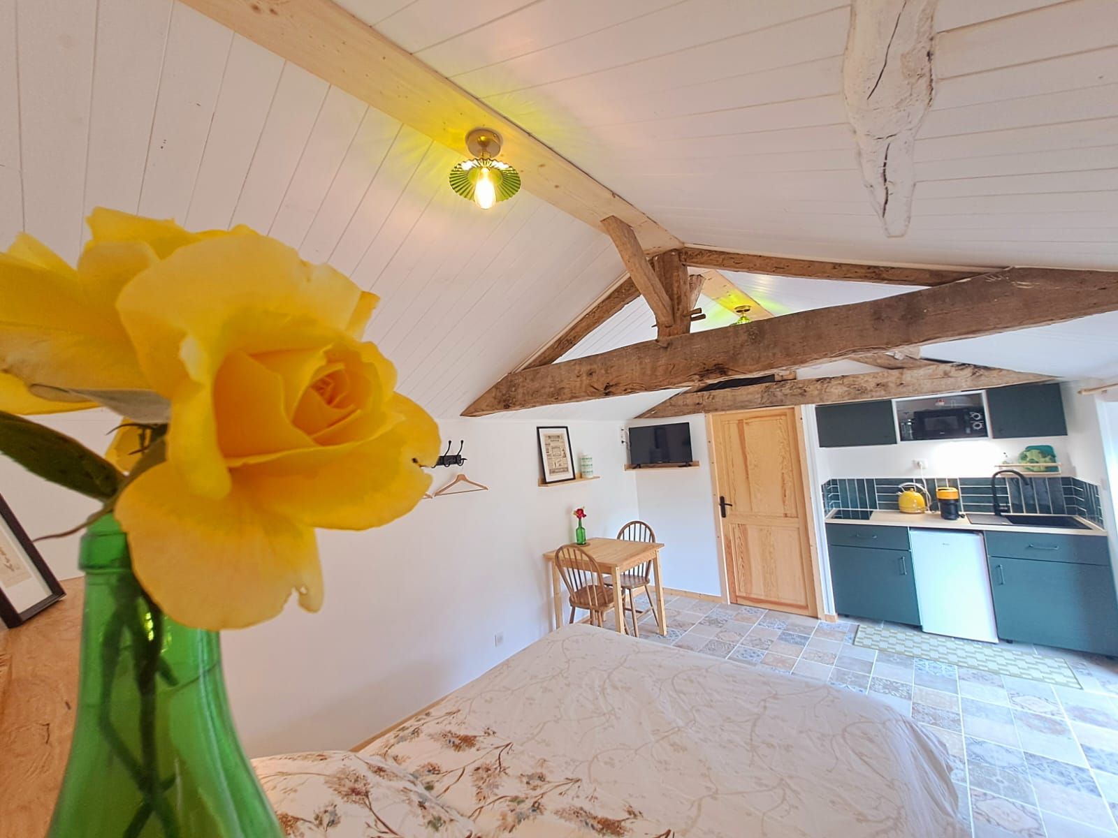 Een kamer met een bed, een houten plafond, een groene vaas met een gele bloem, een kleine tafel en een kitchenette.