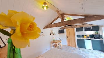 Een kamer met een bed, een houten plafond, een groene vaas met een gele bloem, een kleine tafel en een kitchenette.
