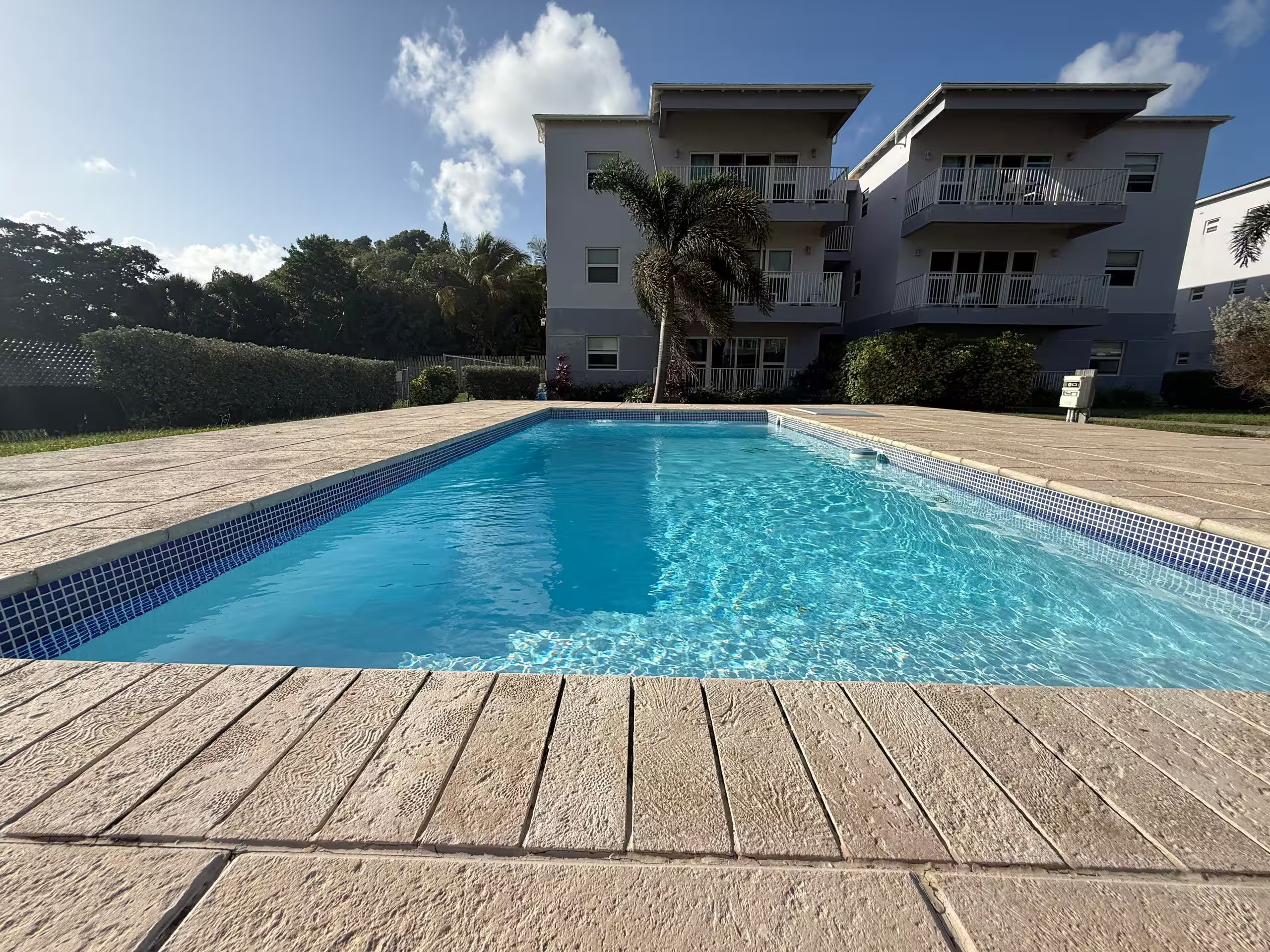 Una piscina rettangolare con bordo piastrellato, circondata da un pontile in legno. Sullo sfondo, un edificio residenziale a più piani con balconi.