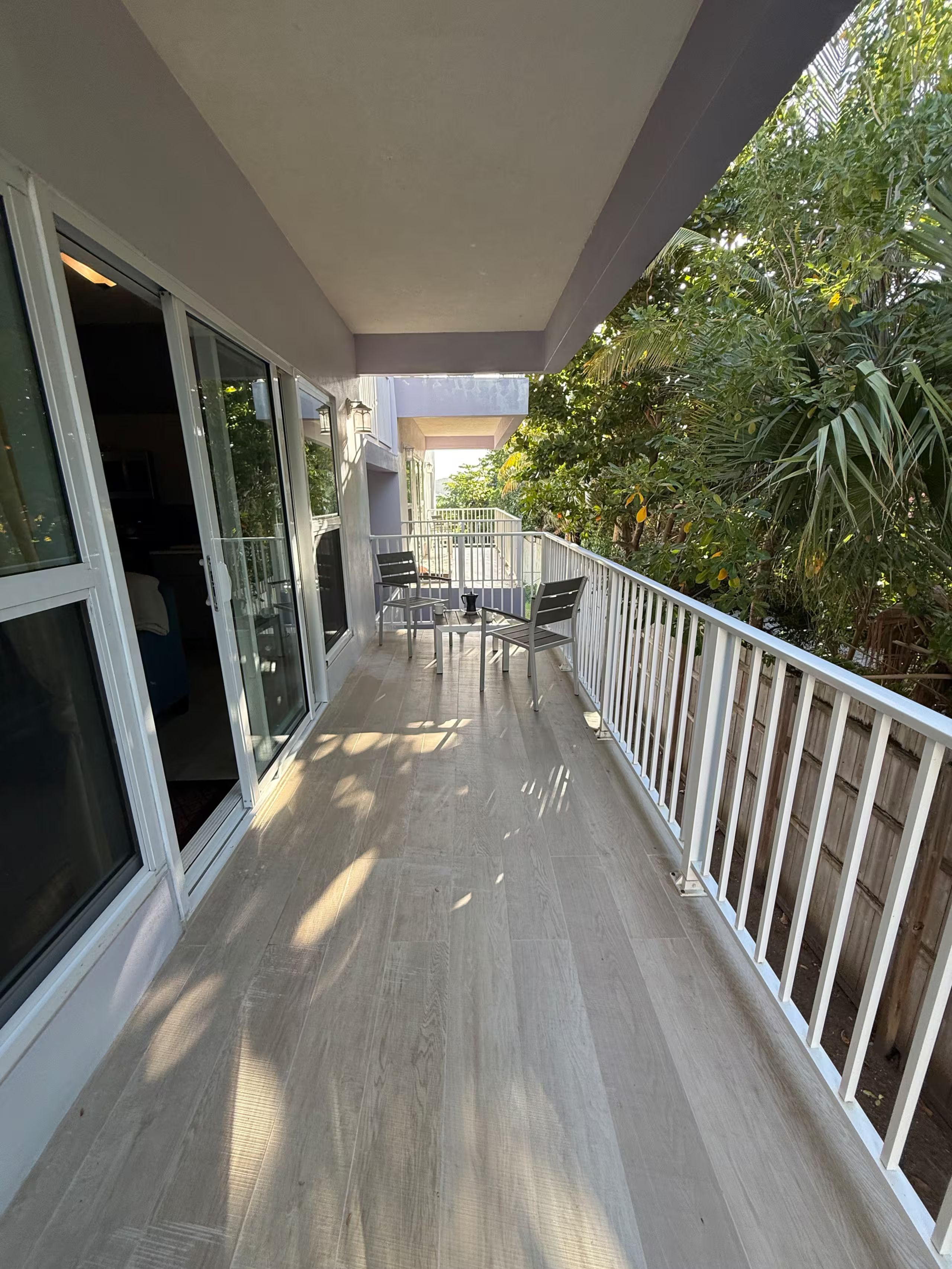 A balcony with a table and chairs, a view of greenery, and a building entrance.
