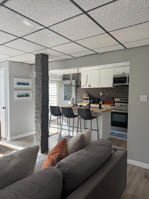A modern kitchen with white cabinets, a microwave, and an oven. There is a bar area with stools and a countertop. - Your Perfect Snowshoe  Getaway make most of the mountains in this stylish condo! (Snowshoe)