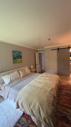 Ein Schlafzimmer mit einem Bett, einem Nachttisch, einer Lampe, einer Tür und einem Flur, der in ein weiteres Zimmer führt.