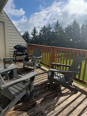 Een terras met Adirondack-stoelen en een barbecue, omringd door een houten omheining en bomen.