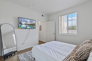 A bedroom with a bed, a TV mounted on the wall, a window with blinds, and a door. - Mesa 2BR Getaway • Near Downtown & Fiesta District (Mesa)