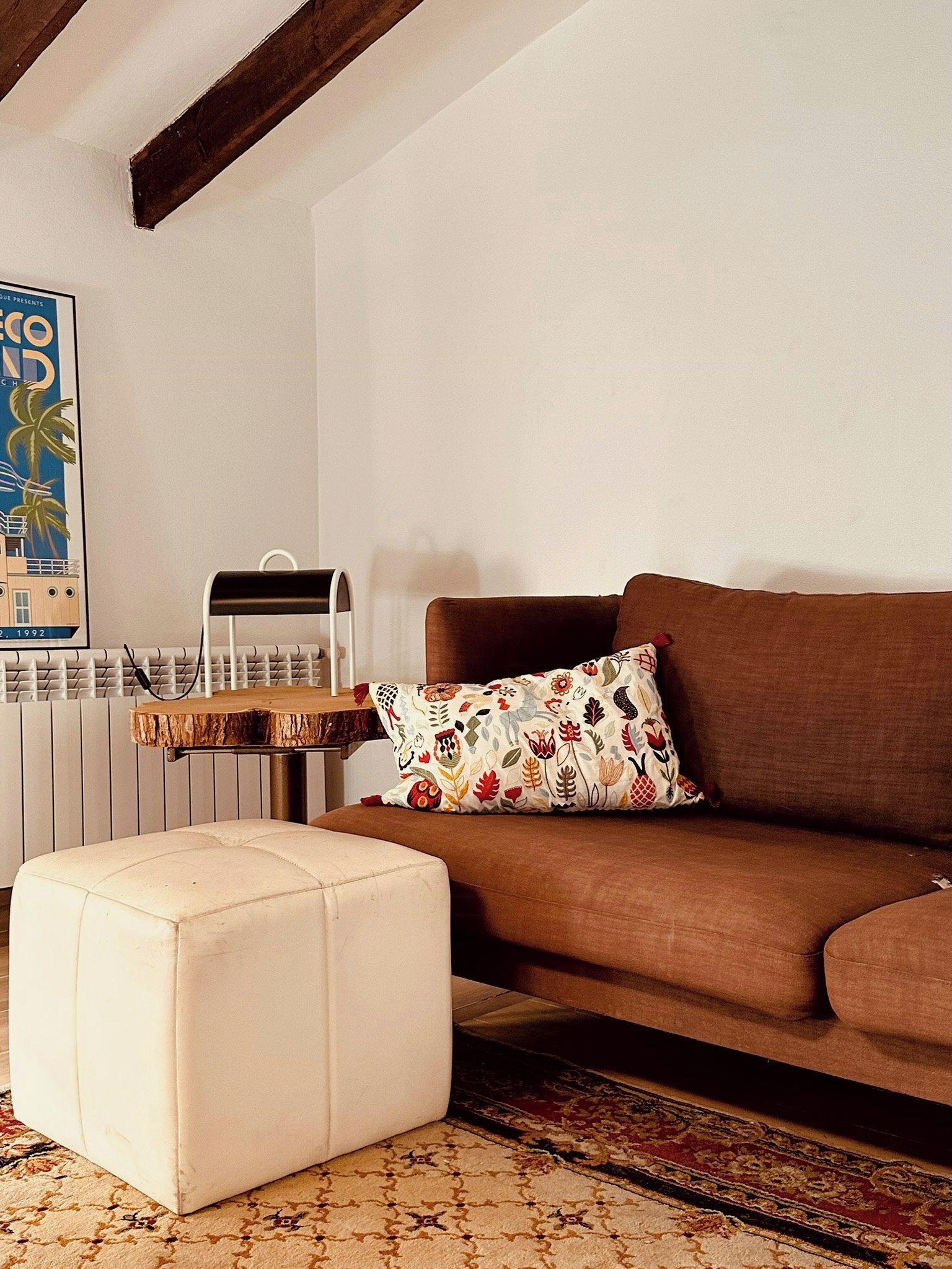 A living room with a brown sofa, a white ottoman, a patterned throw pillow, a wooden coffee table, and a poster on the wall.