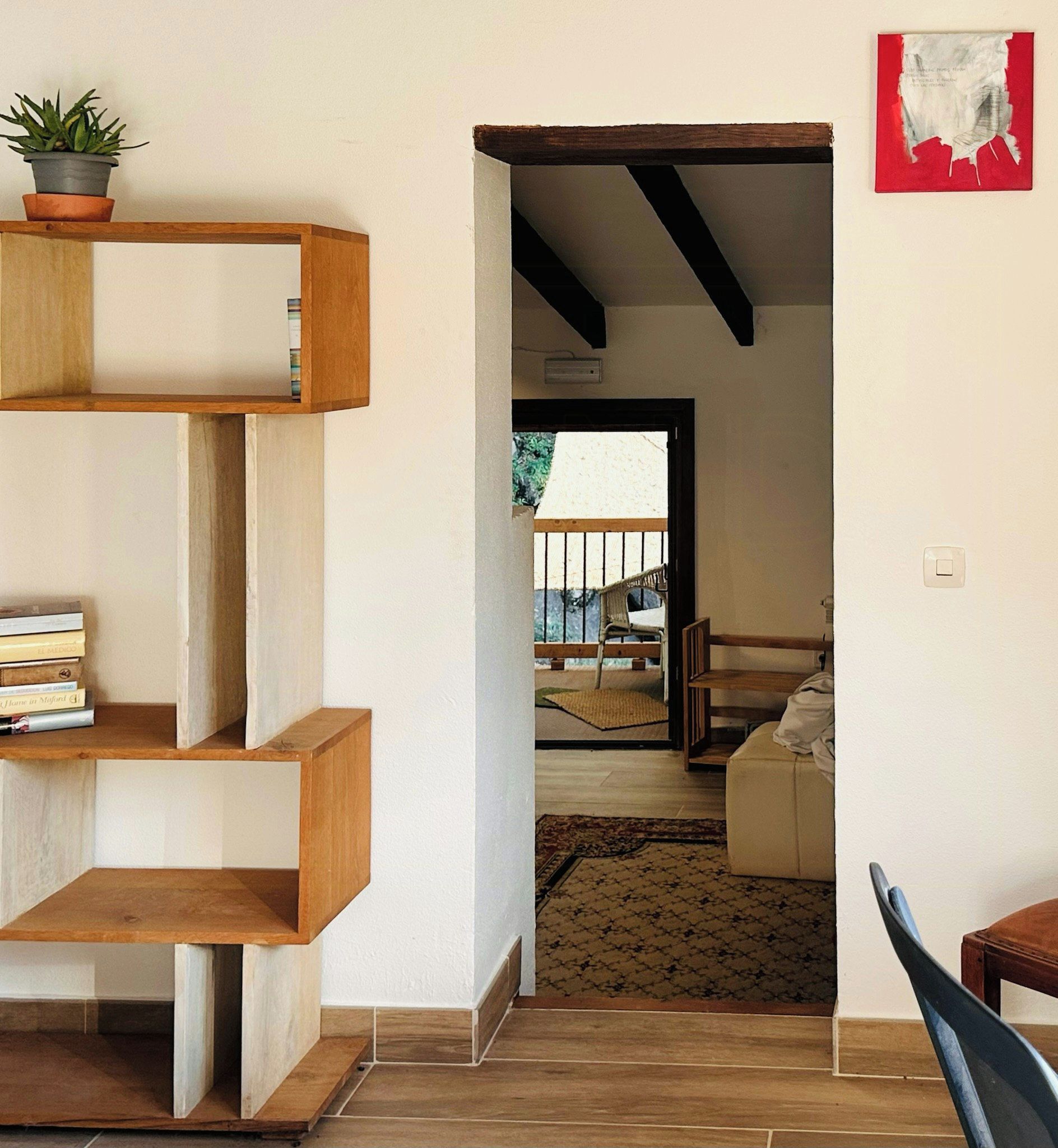 A room with a wooden staircase, a bookshelf, a potted plant, and a view of an outdoor balcony.
