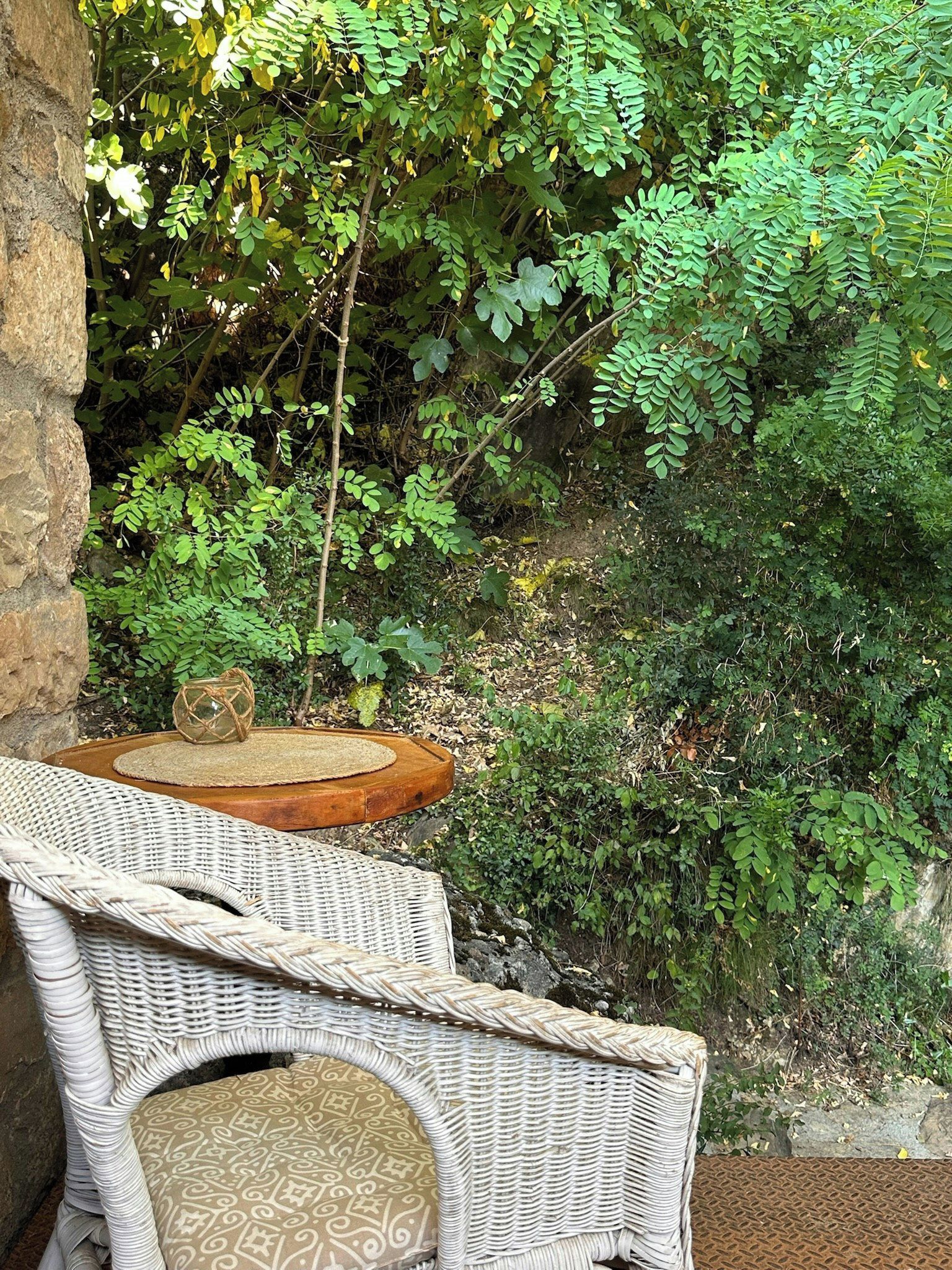 A wicker chair with a patterned cushion, a small wooden table, and a stone wall.