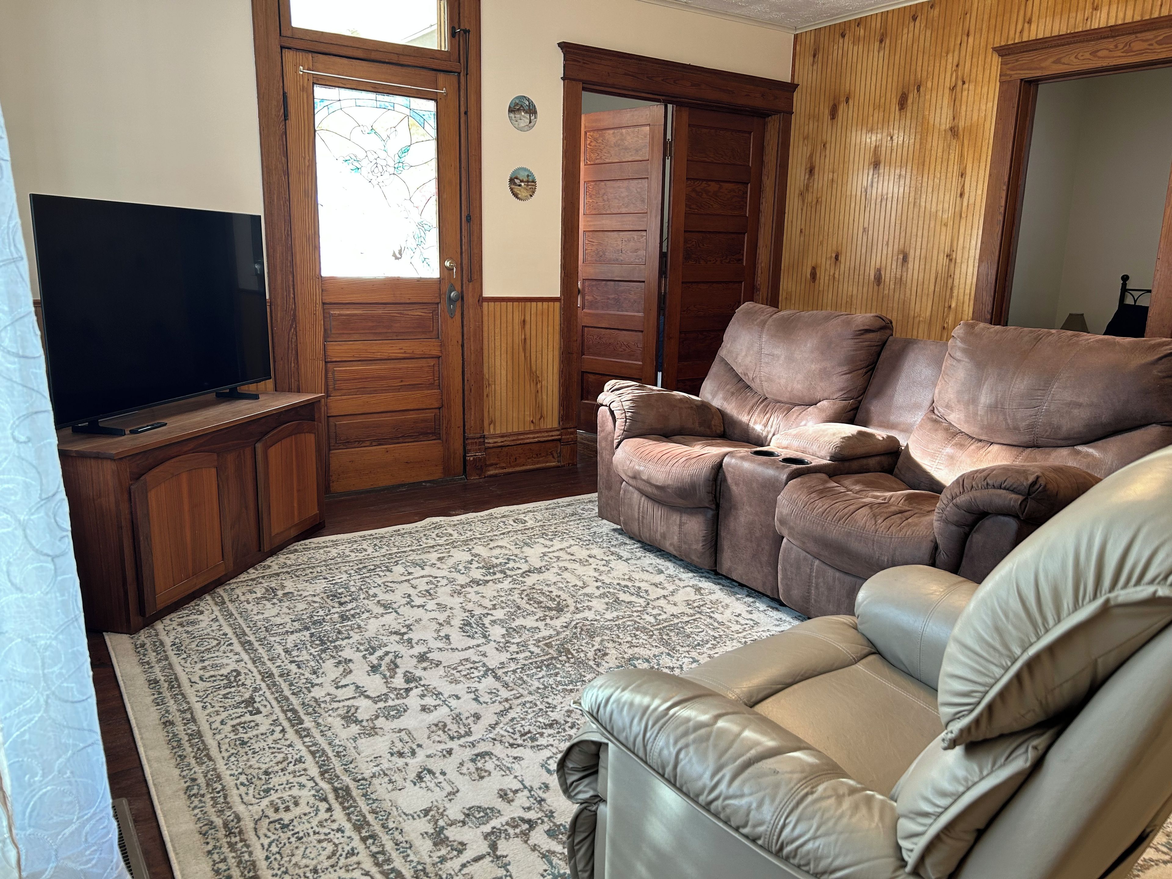 Ein Wohnzimmer mit Fernseher, zwei Sofas, einem gemusterten Teppich und Holztüren.