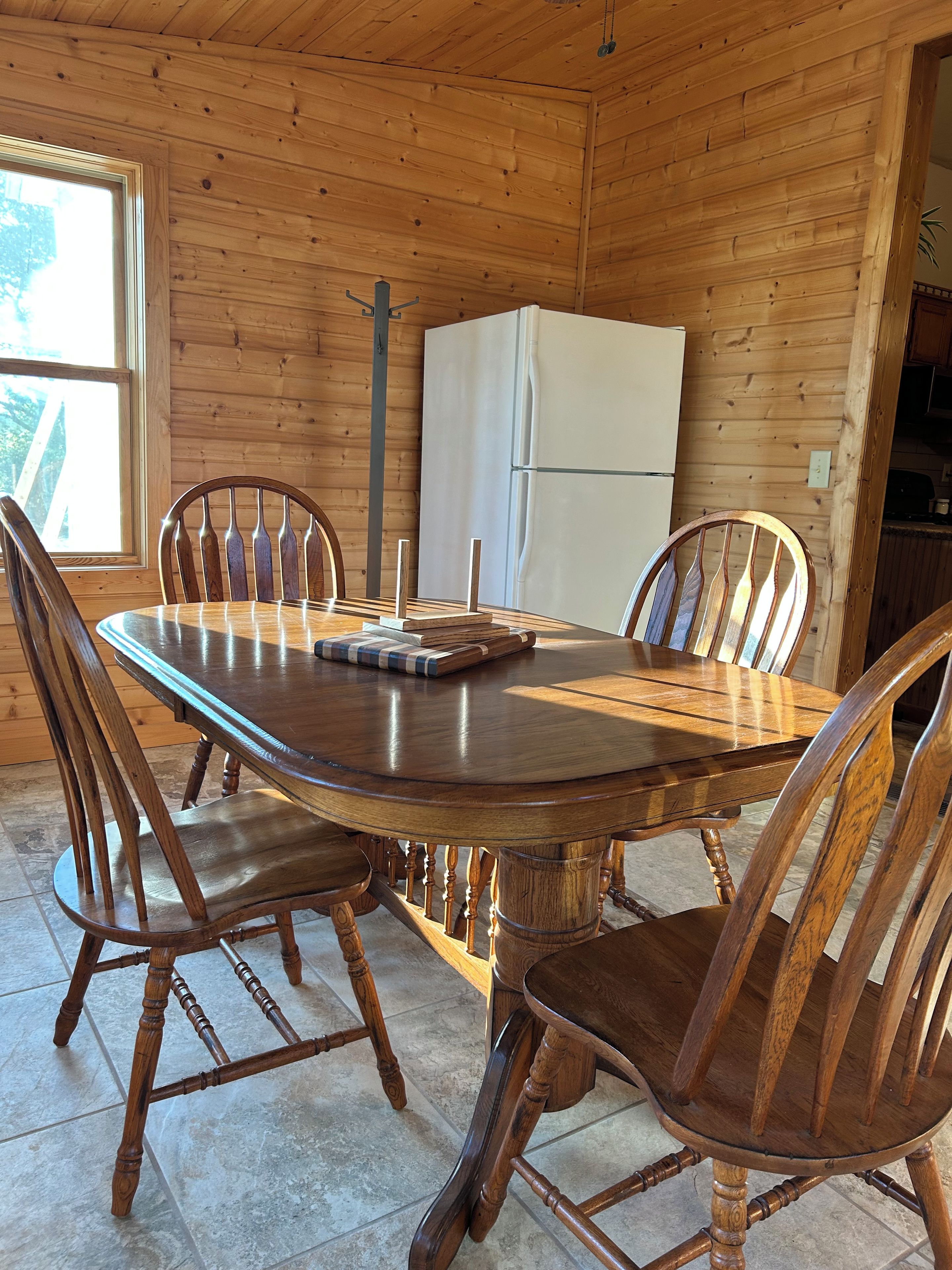 A wooden-paneled room with a round dining table and chairs, a white refrigerator, and a window with a view.