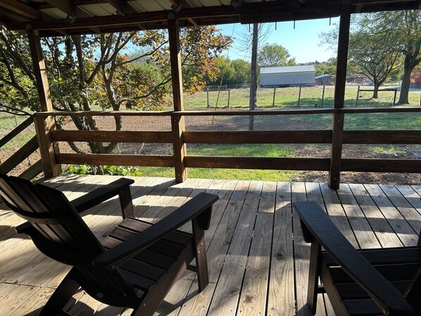 Eine Holzterrasse mit Bänken und einem Tisch, mit Blick auf einen umzäunten Garten mit Bäumen und einem Gebäude im Hintergrund.