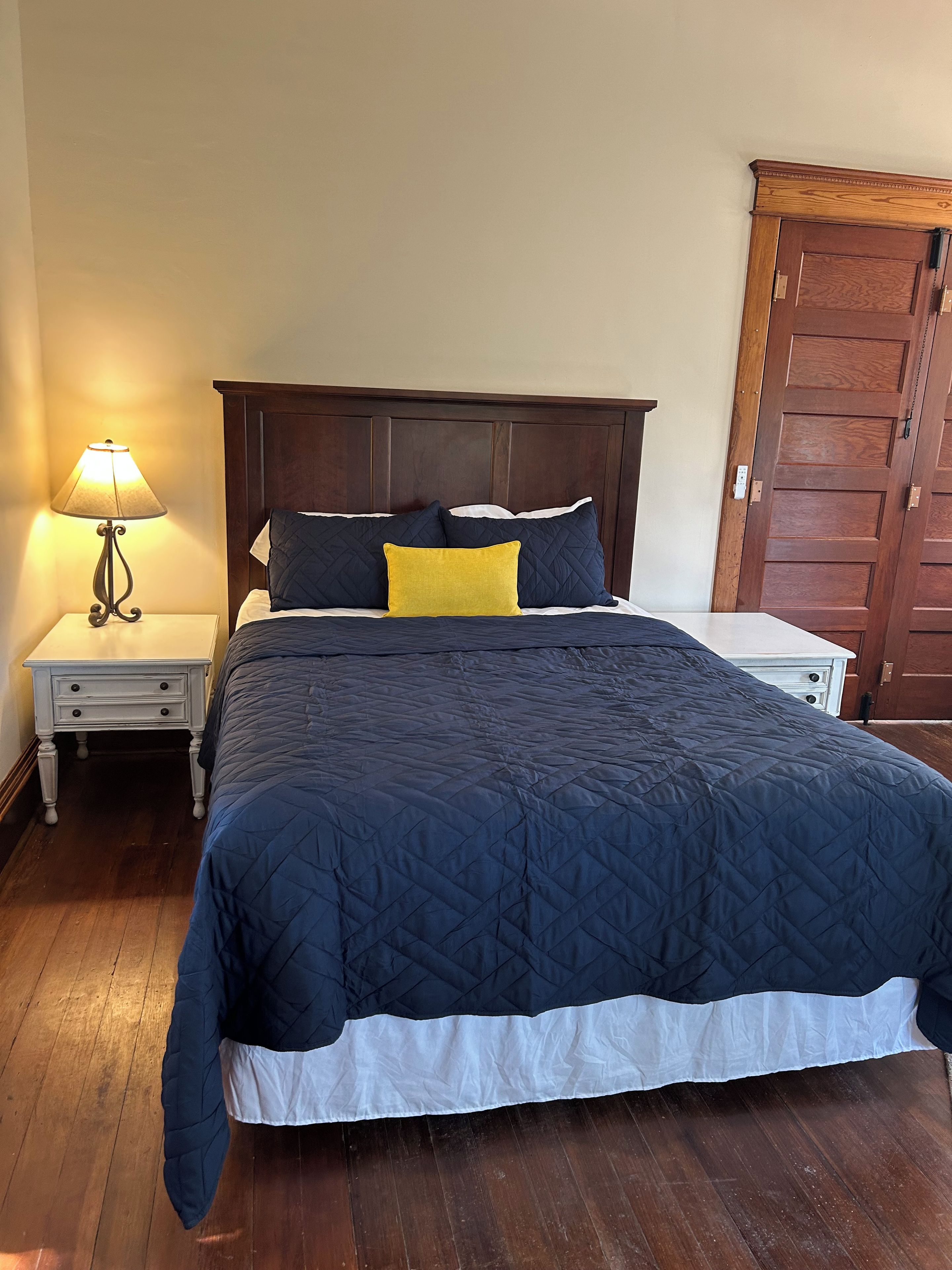Een bed met een donkere gewatteerde spreideken, een bedlampje en een houten garderobe.
