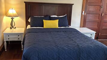 Een bed met een donkere gewatteerde spreideken, een bedlampje en een houten garderobe.