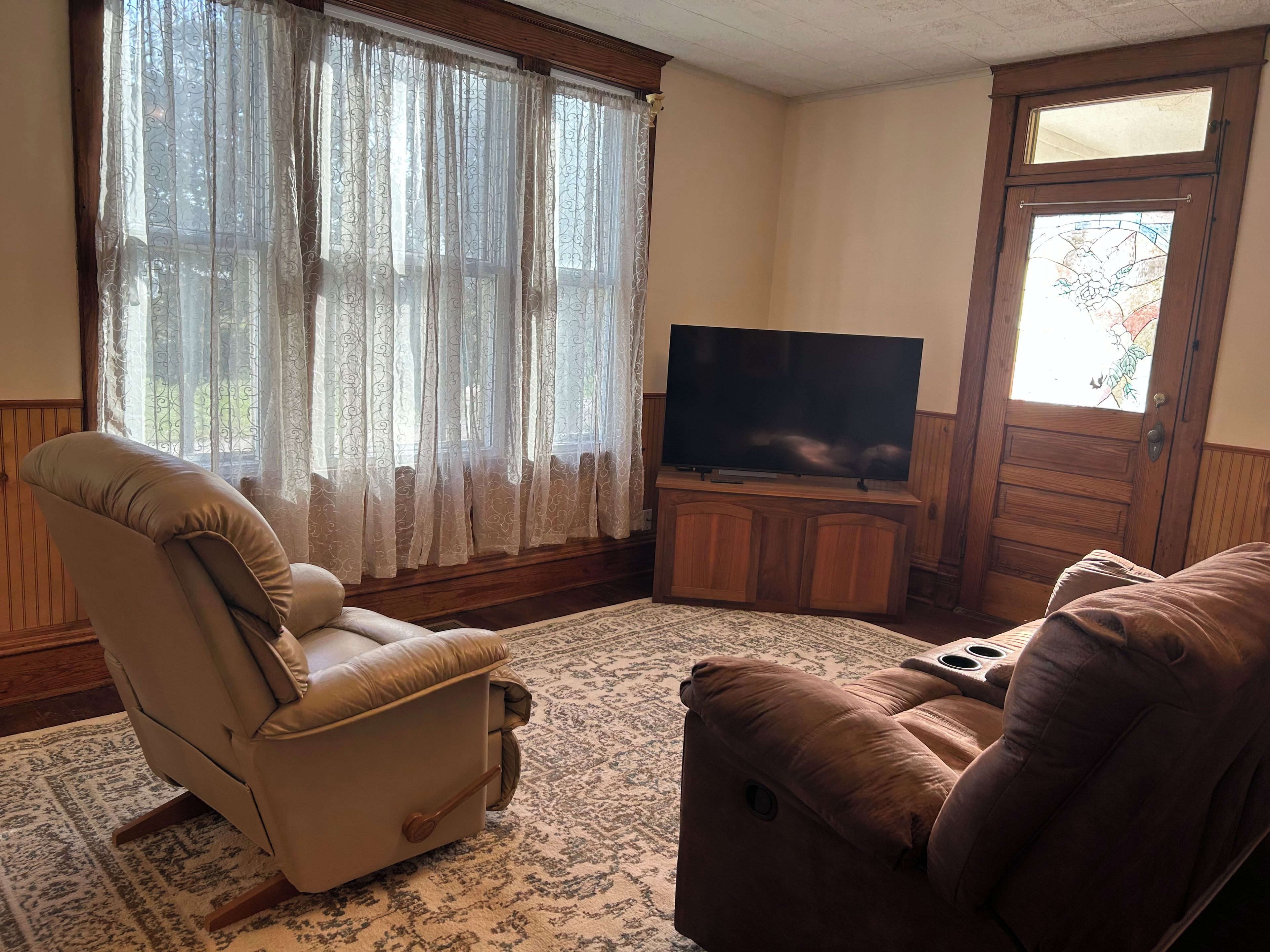 Un salon avec un fauteuil beige, un canapé marron, un téléviseur à écran plat posé sur un meuble en bois et un tapis à motifs.
