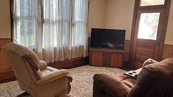 Un salon avec un fauteuil beige, un canapé marron, un téléviseur à écran plat posé sur un meuble en bois et un tapis à motifs.