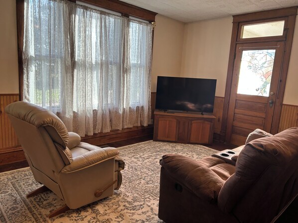 Un salon avec un fauteuil beige, un canapé marron, un téléviseur à écran plat posé sur un meuble en bois et un tapis à motifs.