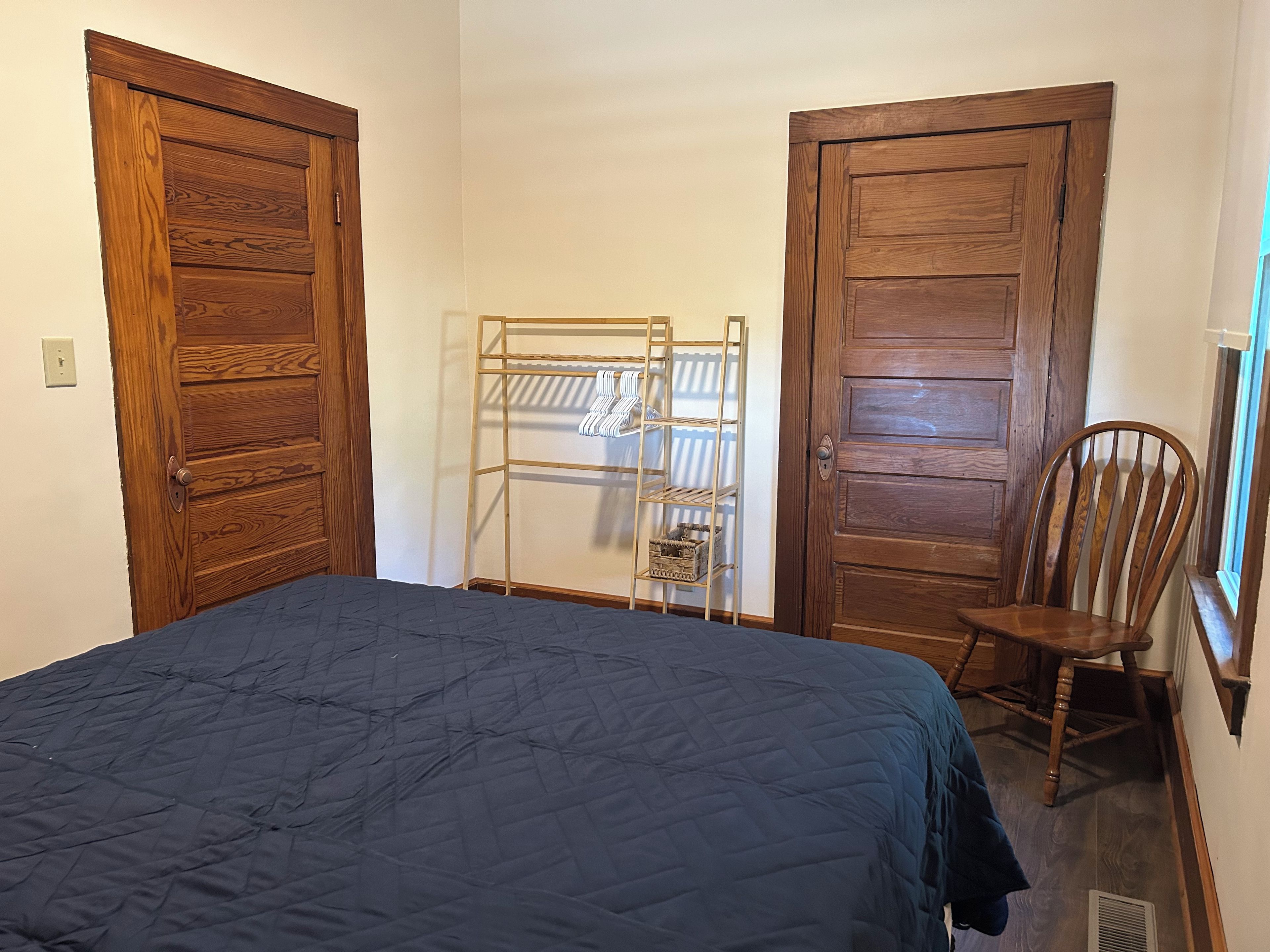 A bedroom with a bed, wooden wardrobe, a metal drying rack, and a wooden chair.