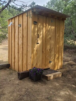 A wooden shed with a door and a small step leading up to it, surrounded by gravel and a planter with purple flowers.