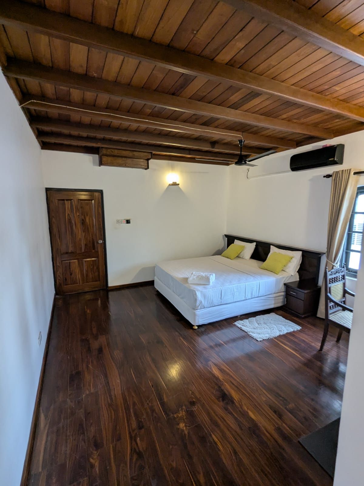 Une chambre avec un plafond en bois, un lit avec une literie blanche et des oreillers jaunes, une table de chevet, une chaise et une fenêtre avec des rideaux.