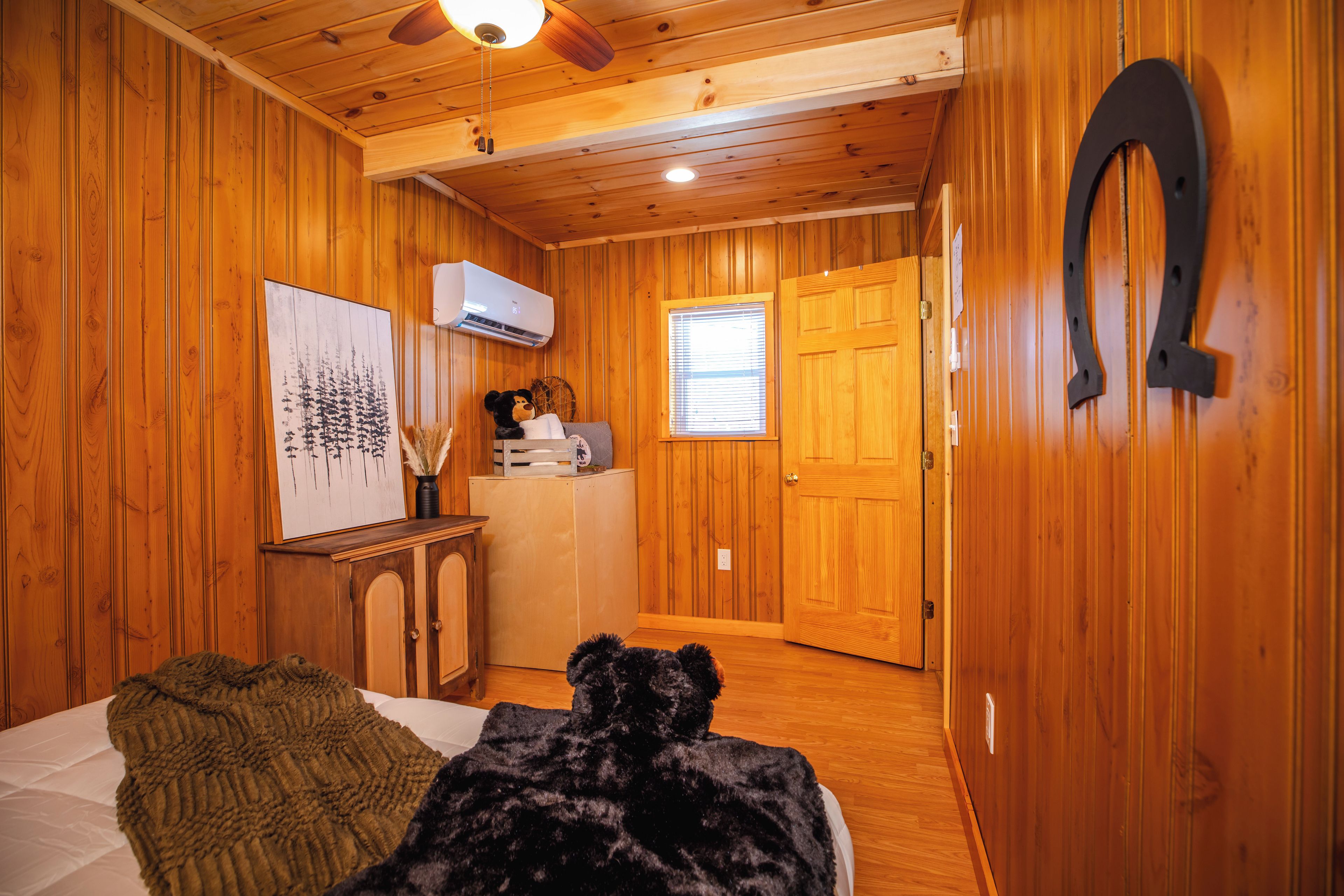 A cozy cabin interior with wooden walls, a ceiling fan, a bed with a green blanket, a black dog, and a wooden door.