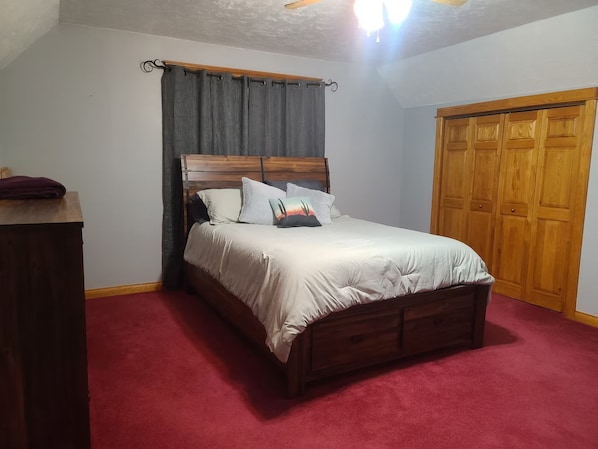 A bedroom with a wooden bed, a red carpet, a wooden wardrobe, and a ceiling fan.