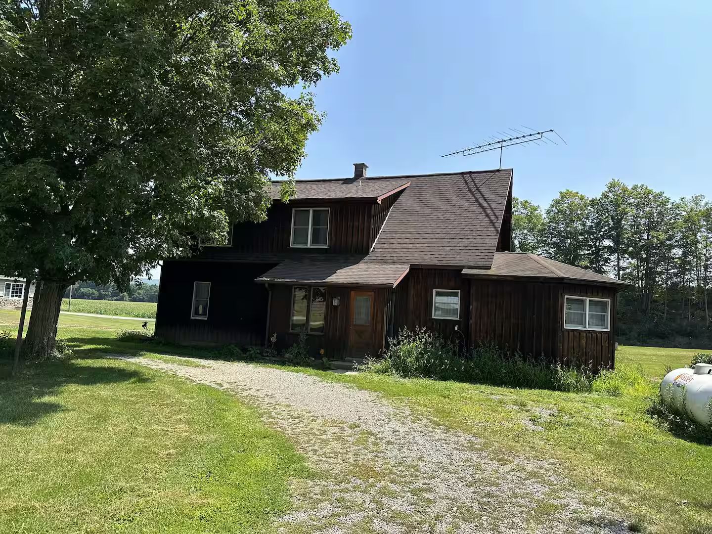 A two-story wooden house with a sloped roof, a gravel driveway, and a large tree in the foreground.