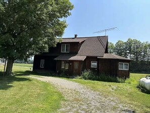A two-story wooden house with a sloped roof, a gravel driveway, and a large tree in the foreground.