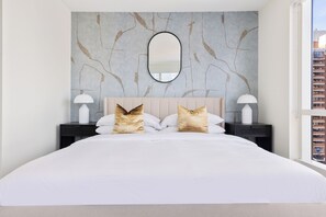 Ein modernes Schlafzimmer mit einem großen Bett, zwei Nachttischen, einem Spiegel und Blick auf Gebäude durch ein Fenster.