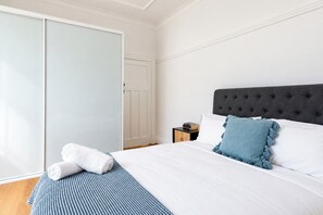 Una cama bien tendida con ropa blanca y una almohada azul decorativa, una mesita de noche con libros, y una puerta cerrada con interruptor de luz.