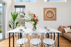 Un comedor con una mesa blanca dispuesta para cuatro personas, rodeada de sillas blancas, un florero con flores y una obra de arte enmarcada en la pared.