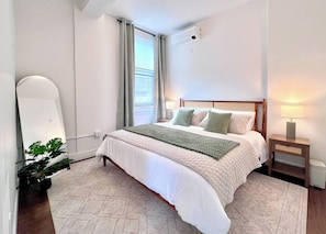 Ein Schlafzimmer mit Bett, Nachttisch, Lampe, Klimaanlage, Fenster mit Vorhängen und einer Zimmerpflanze.