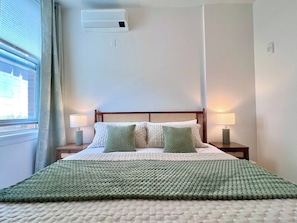 A hotel room with a bed, two bedside lamps, a window with curtains, and an air conditioning unit on the wall. - DWTN New Haven | King Bed | Free Parking | Gym (New Haven)