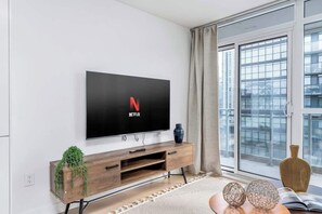 A modern living room with a flat-screen TV mounted on a wall, a wooden entertainment unit with a plant, and a large window with a view of buildings. - Luxe Downtown Suite Steps to Entertainment District (Toronto)