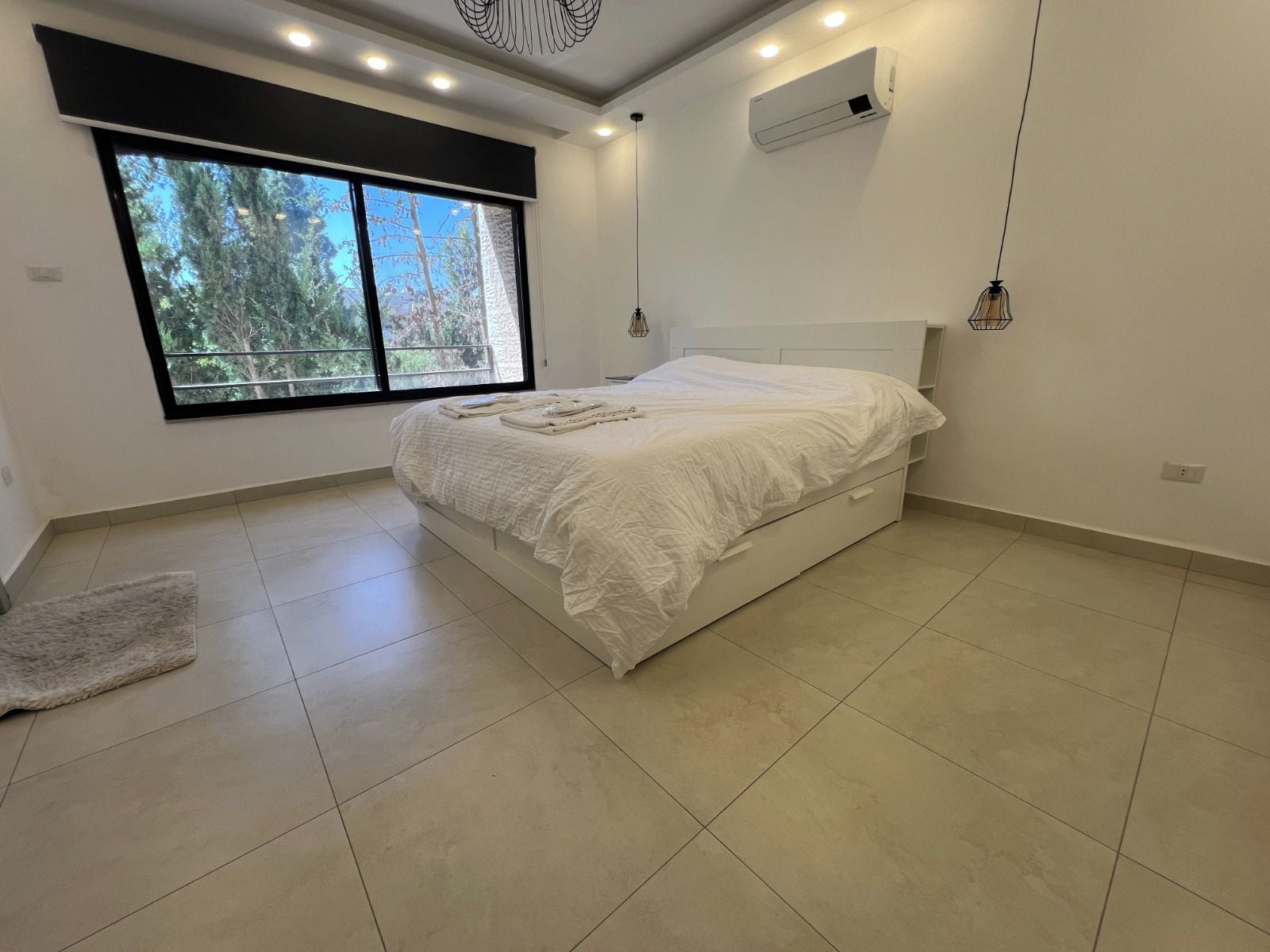 A bedroom with a large window, a bed, a white air conditioning unit, and a ceiling light fixture.