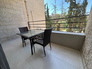 A balcony with a glass table and chairs, a view of trees, and a building in the background. - Cozy 2-Bedroom Apartment with 3 Beds & 2 Bathrooms (Amman)