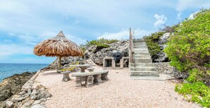 A beach setting with a thatched umbrella, a stone bench, a staircase, and a rocky backdrop. - Villa Azure Horizon - Oceanfront & Private Pool - Coral Estate Resort - Privacy (Coral Estate)