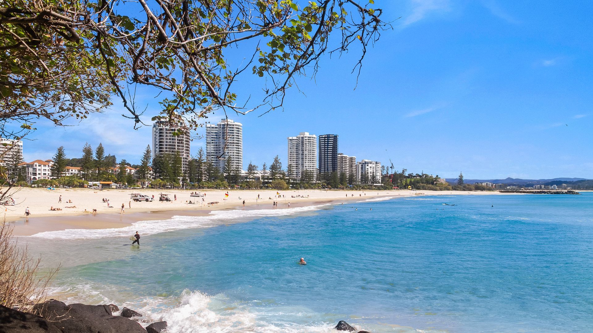 A beach with clear blue waters, a sandy shore, and tall buildings in the background.
