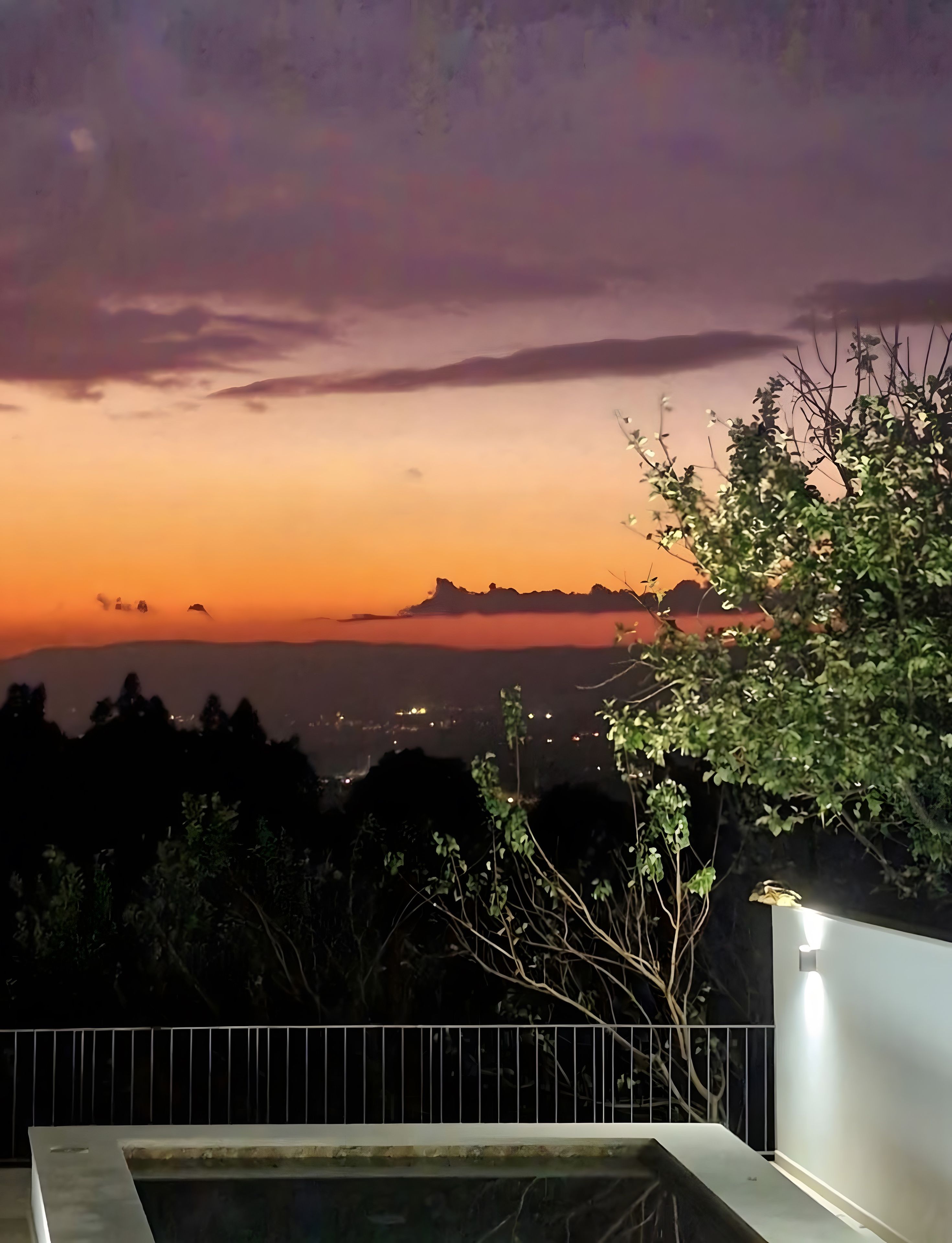 A nighttime view from a balcony with a railing, overlooking a cityscape with distant mountains and a sunset sky.